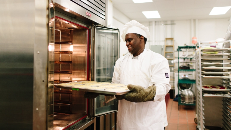 A chef from Hot Bread Kitchen wearing a white uniform and hat places a tray of food into a commercial oven. He is in a professional kitchen with industrial equipment and trays on racks in the background. Photo by Christine Han, courtesy of Hot Bread Kitchen. A chef from Hot Bread Kitchen wearing a white uniform and hat places a tray of food into a commercial oven. He is in a professional kitchen with industrial equipment and trays on racks in the background. Photo by Christine Han, courtesy of Hot Bread Kitchen.