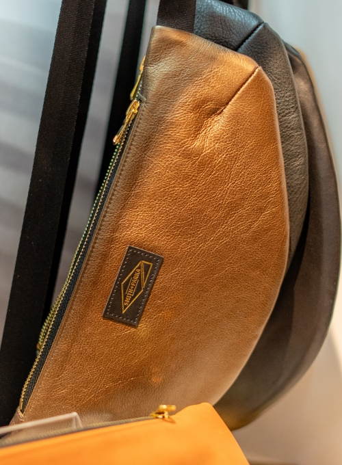 Close-up of a stylish sling bag with a metallic gold and black design. The bag features a diagonal zipper and a rectangular brand label. A soft focus hints at other bags in the background. Close-up of a stylish sling bag with a metallic gold and black design. The bag features a diagonal zipper and a rectangular brand label. A soft focus hints at other bags in the background.